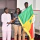 Three people on a stage; a flag with green, yellow, and red is being held aloft during a formal ceremony.