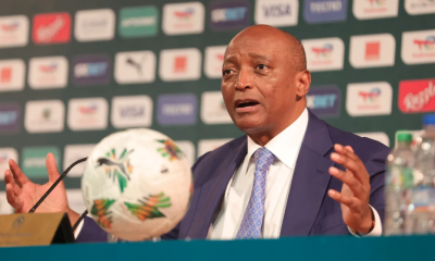 Man in a blue suit and light blue tie speaks at a press conference with a football on the table nearby.