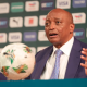 Man in a blue suit and light blue tie speaks at a press conference with a football on the table nearby.