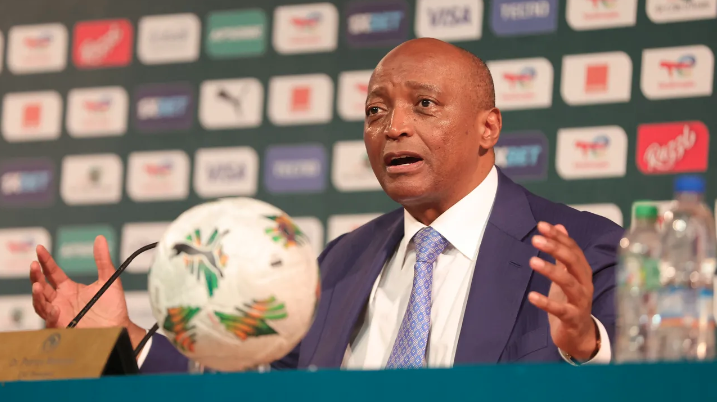 Man in a blue suit and light blue tie speaks at a press conference with a football on the table nearby.