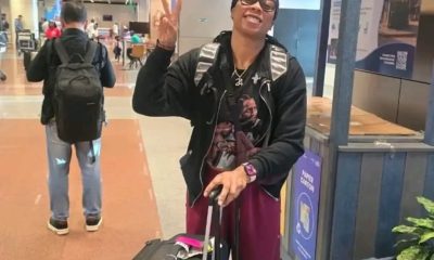 Smiling traveler in a black jacket and glasses waves a peace sign while pulling a black rolling suitcase at the airport.