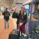 Smiling traveler in a black jacket and glasses waves a peace sign while pulling a black rolling suitcase at the airport.