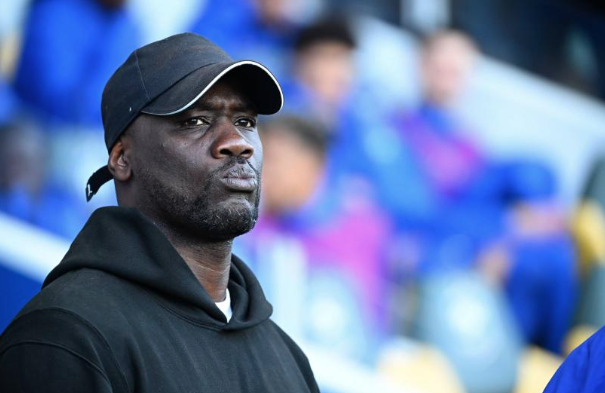 Man in a black hoodie and cap stands in a stadium, looking off to the side with a focused expression.