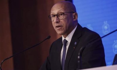 Bald man wearing glasses in a dark suit speaks at a podium with a microphone during a conference, blue backdrop behind him.