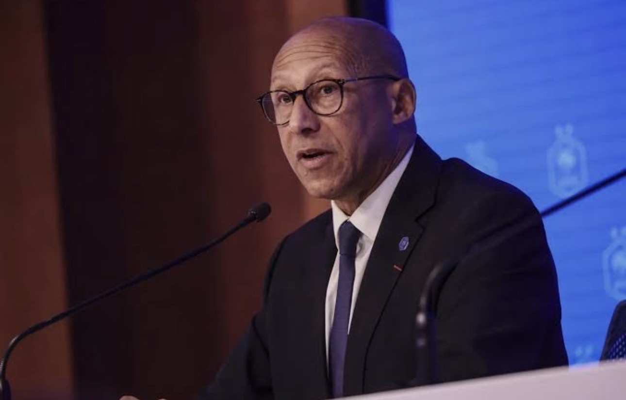 Bald man wearing glasses in a dark suit speaks at a podium with a microphone during a conference, blue backdrop behind him.